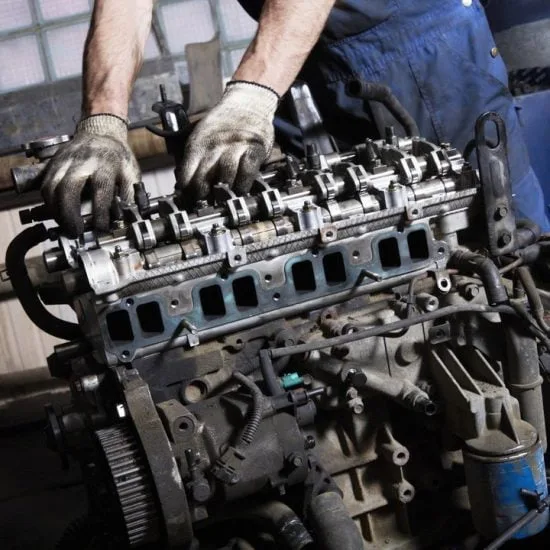 A person wearing gloves is working on an engine in a workshop, focusing on components and mechanical details, surrounded by tools.