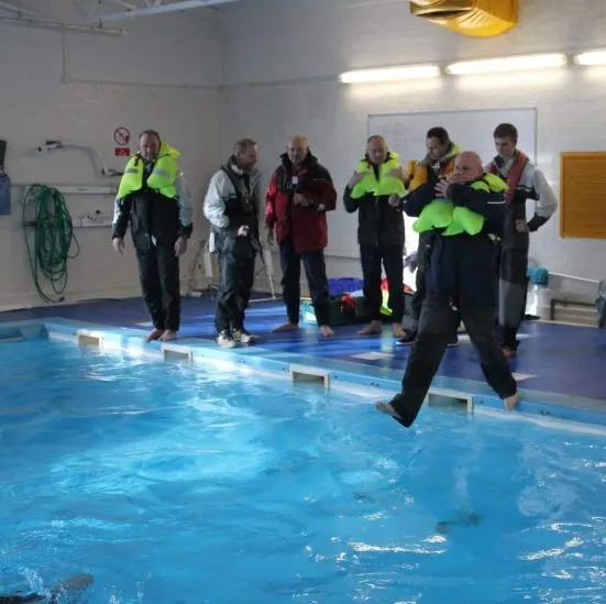 People wearing life jackets stand by an indoor pool. One person steps into the water, suggesting a safety or survival training exercise.