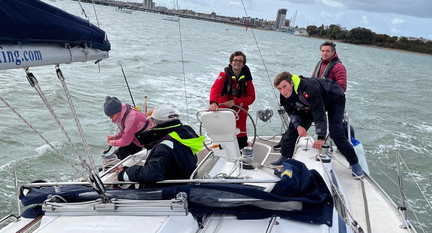 Five people in sailing gear on a yacht, navigating a bay with distant shoreline and cityscape, including modern buildings and docks.