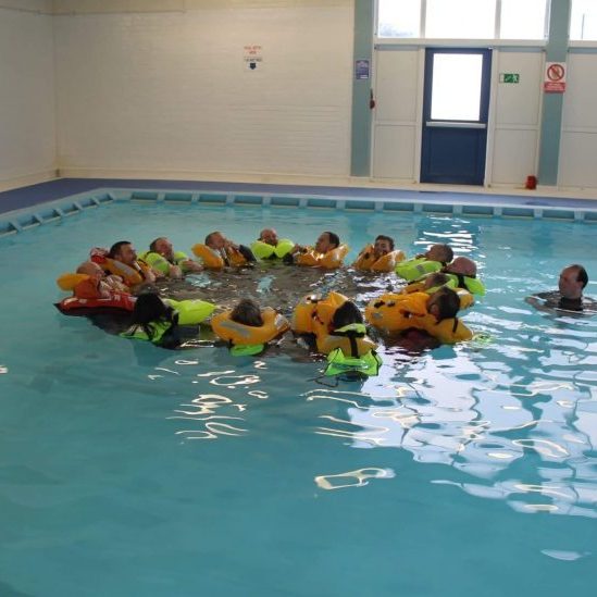 A group of people in lifejackets forms a circle in a swimming pool, participating in water safety training indoors.