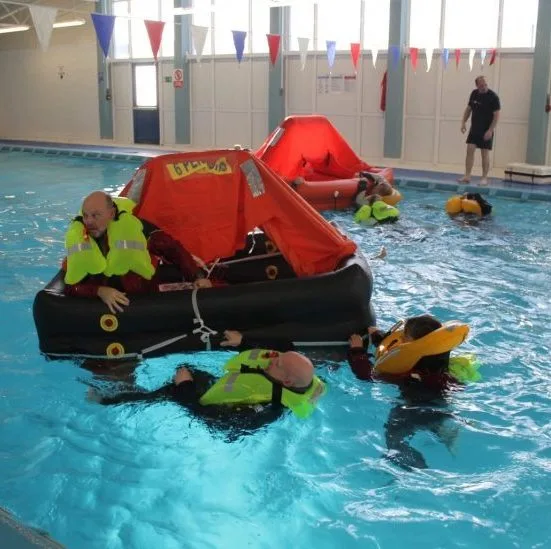 People in life jackets participate in a survival training drill in a swimming pool, using inflatable rafts and gear for practice.