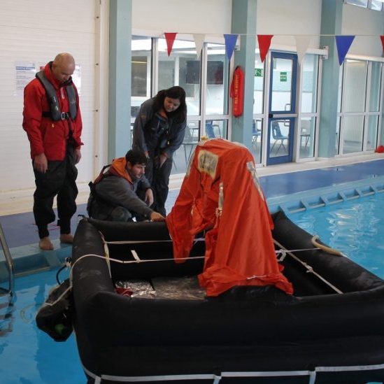Three people inspect an orange life raft in an indoor pool area. Safety equipment and colorful flags are visible in the background.
