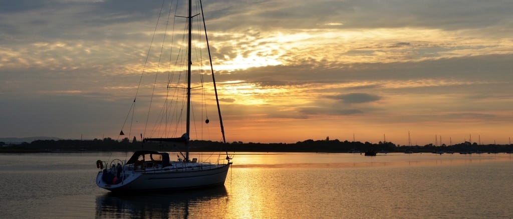 A sailboat floats on calm water at sunset, with a serene sky and distant shoreline, creating a peaceful, picturesque scene.