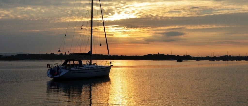 A sailboat floats on calm waters during a peaceful sunset, with silhouetted shoreline and sky displaying shades of orange and gold.