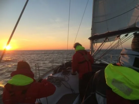 Three people in bright jackets sailing on open water at sunset, navigating a boat's deck under a colorful sky.