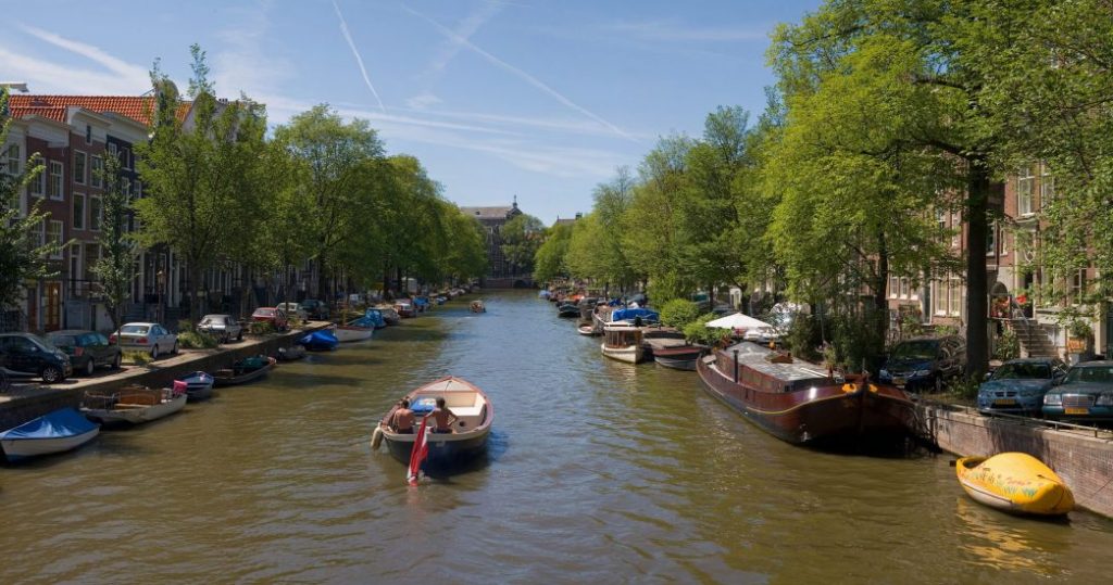 A person navigates a boat down Amsterdam's tree-lined canal, flanked by historic houses and parked cars under a clear blue sky.