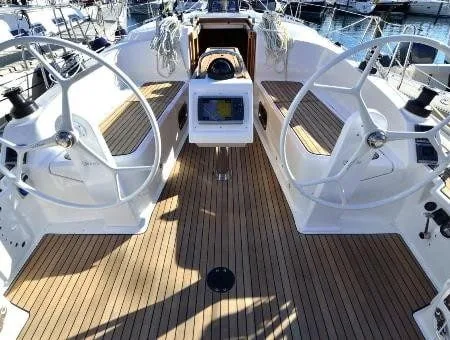 View from a sailboat's cockpit with dual steering wheels, a central navigational screen, and a wooden deck, docked in a marina.