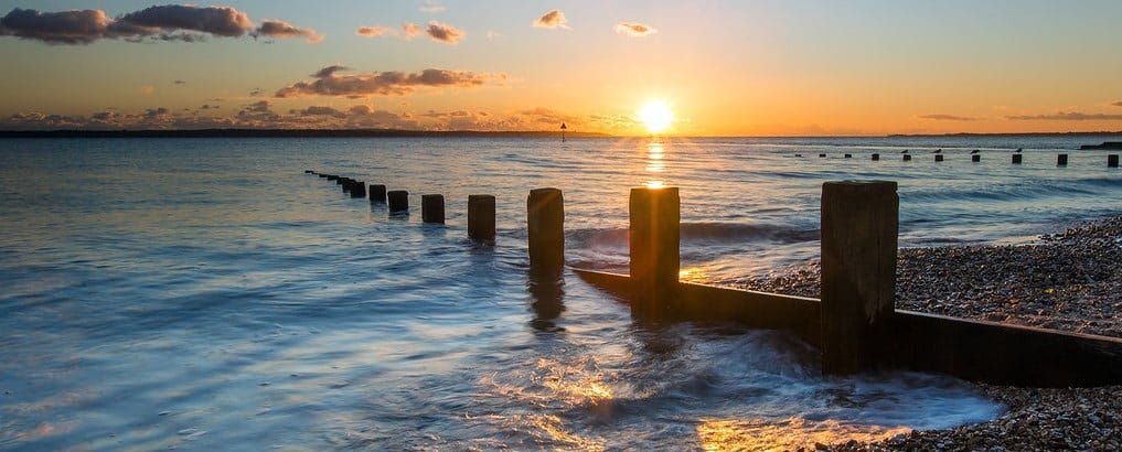 A serene sunset over the ocean with wooden groynes stretching into the water and pebble-laden shore, under a softly lit sky.