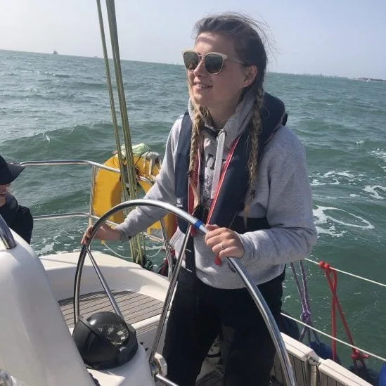 A person steering a sailboat on the ocean, wearing sunglasses and a life jacket, with a clear sky and distant ships visible.