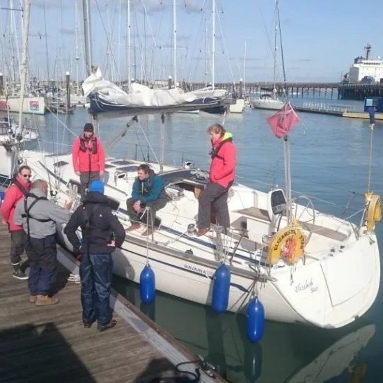 Five people gather on a yacht docked at a marina. Sailboats and a pier are visible in the background under a clear sky.