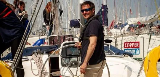 A person smiles on a sailboat, surrounded by equipment and other boats at a marina. "Clipper" sign visible in the background.