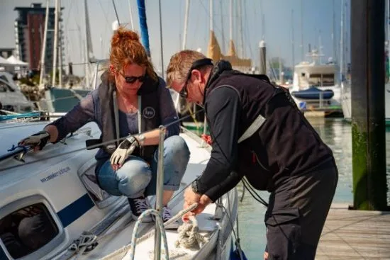 Two people working on a sailboat in a marina, surrounded by other boats on a sunny day, tying ropes and wearing life vests.