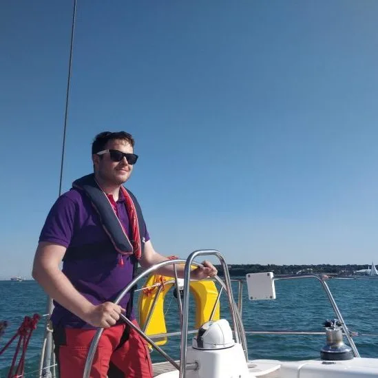 A person steering a sailboat on clear blue water, wearing sunglasses and a life jacket, with a sunny sky overhead.