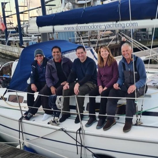 Five people sitting on a docked yacht at a marina, ropes visible. The boat displays a website at the top. Overcast sky background.