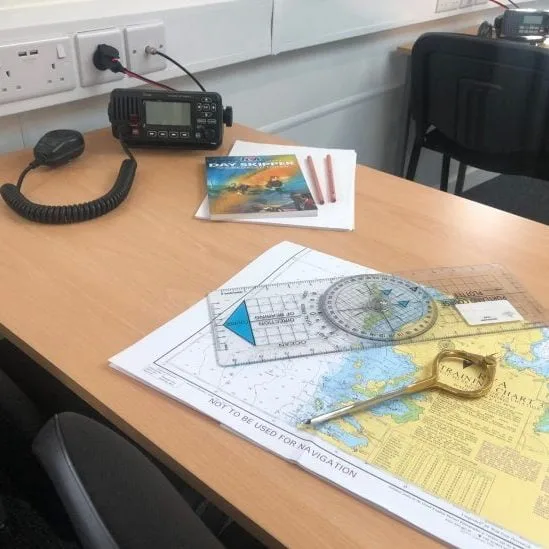 A desk displays a marine radio, navigation map, protractor, ruler, pencils, and book, creating a nautical-themed setup for maritime navigation study.