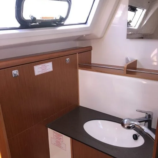 A compact boat bathroom features a small sink, shiny faucet, wooden cabinetry, and a porthole window, providing natural light and a nautical feel.