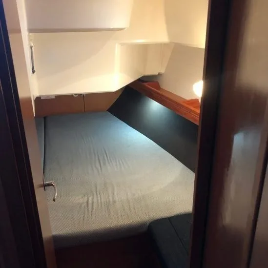 A compact bedroom on a boat features a bed with blue sheets, wooden trim, white walls, shelving, and cozy lighting.