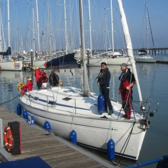 Five people stand on a docked sailboat in a marina, surrounded by other boats on a clear, sunny day.