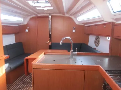 Interior of a sailboat featuring wooden furnishings, a compact kitchen with stainless steel fixtures, and cozy seating area beneath large windows providing natural light.