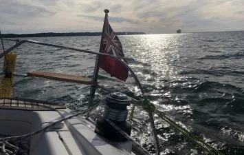 Sailboat on a sunlit sea with a flag at the stern. A distant shoreline and ship are visible, great day in the solent.