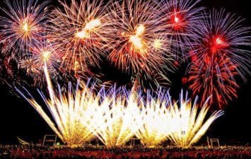 A vibrant fireworks display illuminates the night sky above a large crowd, creating a spectacular burst of colors and light patterns.