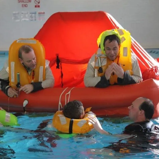Four people in life vests practice survival skills in a pool with an orange life raft. No recognizable landmarks present.