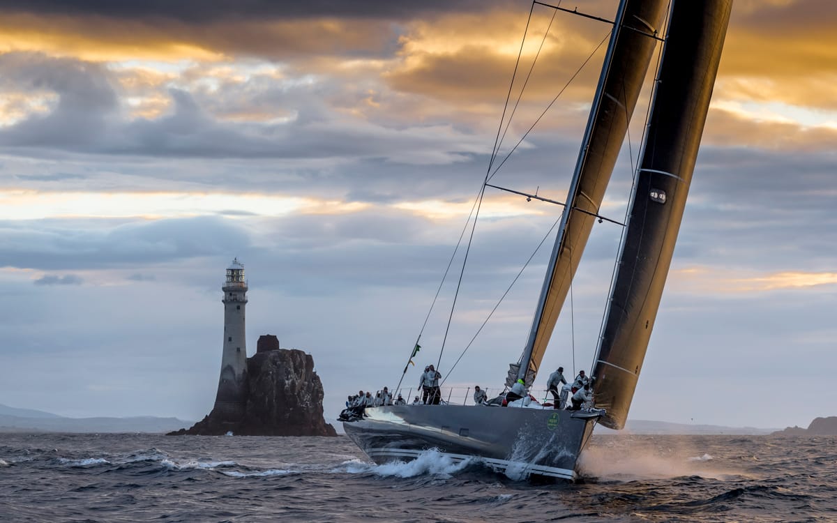 A sailing yacht with several people navigates near Fastnet Rock Lighthouse, under a dramatic sky at sea during sunset or sunrise.