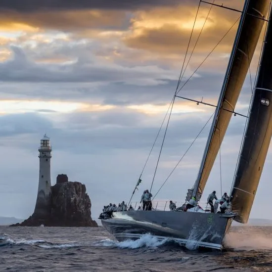 A sailboat with people navigates near Fastnet Rock lighthouse at sunset, with dramatic clouds overhead and waves below.