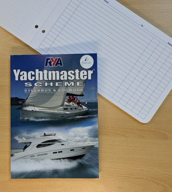 A Yachtmaster Scheme syllabus and logbook on a wooden table beside a blank log sheet with blue lines.