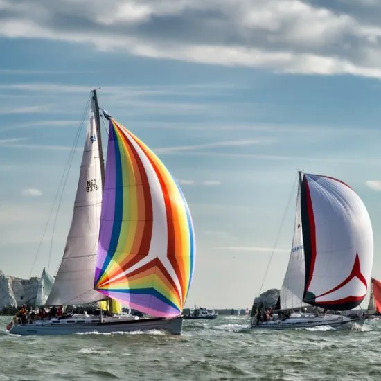 Two sailboats with colorful sails race on the sea under a partly cloudy sky, with multiple people aboard, cliffs in the background.